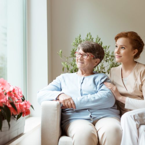 Geriatric woman and her professional caretaker sitting on a couch and looking through a window in a living room. Blooming flowers on a windowsill. Geriatric woman and her professional caretaker sitting on a couch and looking through a window in a living room. Blooming flowers on a windowsill.